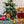 Load image into Gallery viewer, P.L.A.Y. Home for the Holidays Collection Paw Warmers Toy - mittens hanging from a brown dog&#39;s mouth staring into the camera while standing up with a Tree to the left with a present in front and other toys on top of it
