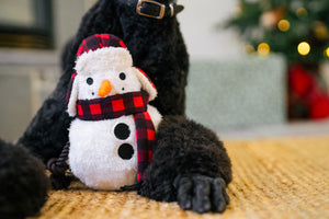 P.L.A.Y. Home for the Holidays Blizzard Buddy Toy - sitting up between two paws of a Black Poodle that is laying down on a woven rug